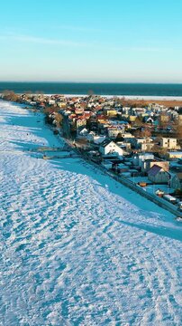 Cinematic vertical drone shot over the snowcovered coast of Rewa, Poland. Features a frozen sea, charming coastal houses, and golden hour light. Perfect for travel and seasonal content.