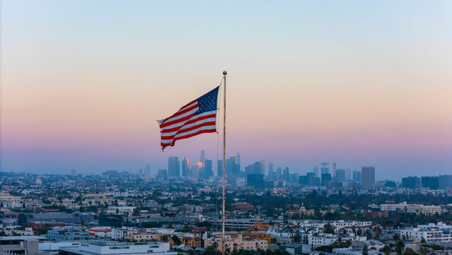 Aerial view of the American flag waving on a flagpole with the Los Angeles skyline in the background during sunset Los Angeles, California, United States.