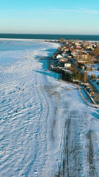 Cinematic vertical drone shot of the snow covered coastline in Rewa, Poland. People walk on the frozen sea ice near a seaside village during a sunny winter morning.