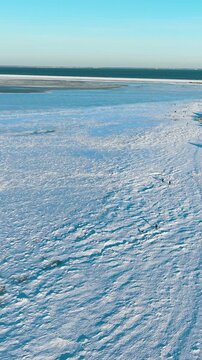 An expansive vertical aerial shot capturing the frozen landscape of Rewa beach during winter. Small figures walk across the ice, highlighting the scale of the cold, bright environment.