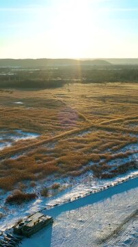 A cinematic vertical drone shot of snowy wetlands and golden reed fields during a bright winter sunset, featuring lens flare and natural textures.