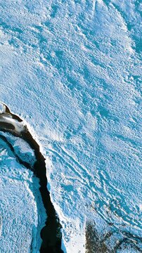 A cinematic vertical aerial view of a dark river cutting through a pristine white snowcovered landscape during a cold winter day. Use for nature documentaries or social media stories.