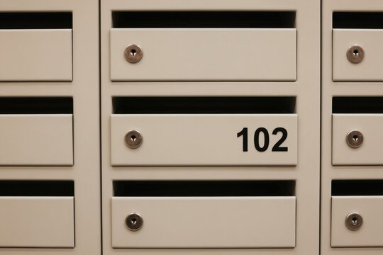 White metal mailboxes in a row for letters and packages. Each secure slot has a lock and some units display apartment numbers. Modern postal delivery system for residents.