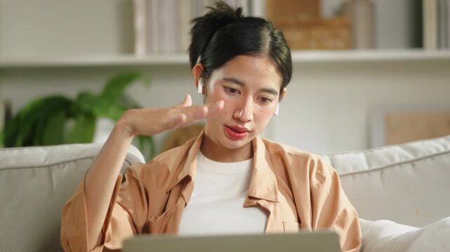 Happy Asian woman using laptop for video meeting on sofa at home, remote working corporate employee, digital nomad hybrid workplace business executive and flexible lifestyle technology concept