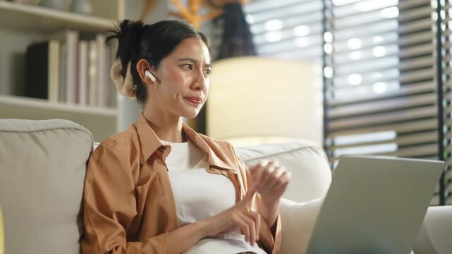 Happy Asian woman using laptop for video meeting on sofa at home, remote working corporate employee, digital nomad hybrid workplace business executive and flexible lifestyle technology concept