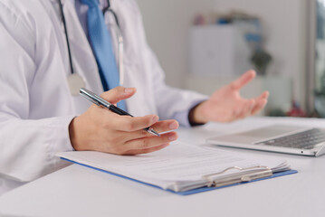 Fototapeta premium Medical consultation and professional advice. Male doctor wearing a stethoscope gesturing while talking at his workplace. Clinic desk with clipboard records and computer.