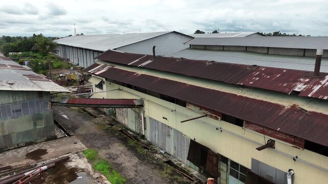An aerial view showing an abandoned factory complex with rusty warehouses, a cluttered scrap metal yard littered with debris, puddles of water, tanks, and newer industrial buildings in the background.