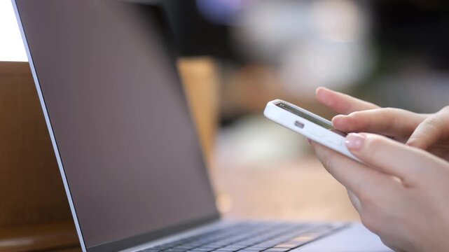 Close-up of hands holding smartphone. Fingers scroll or tap screen actively. Laptop sits nearby, open and unused. Focus sharp on device and hands, background softly blurred. Suggests multitasking