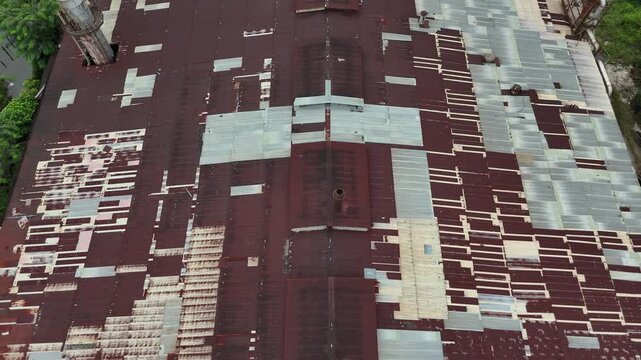 An aerial view captured by a drone showing the roof of a large, abandoned factory, with rusted corrugated metal panels that have been patched up repeatedly, ridgelines, and scattered smokestacks.
