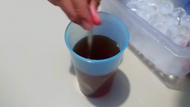 The process of stirring the tea and adding ice cubes to the tea in a blue plastic cup.