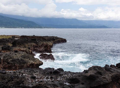 Sanxiantai rocky coastline with waves crashing. Distant mountains under cloudy sky. Taiwan's scenic eastern shore.