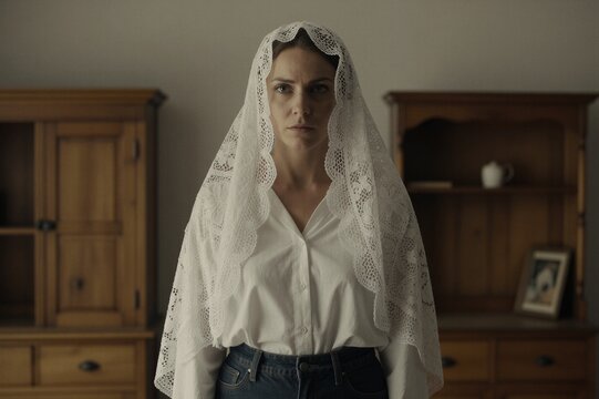 A woman stands serenely in a softly lit room filled with traditional wooden cabinets, her white lace mantilla and button-up shirt a gentle contrast to the warm tones of the rustic