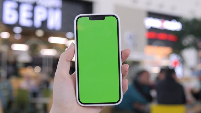 Hand holds smartphone with green screen in busy shopping area. Background shows blurred shoppers and bright signage. Ideal for digital ads or app interface mockups