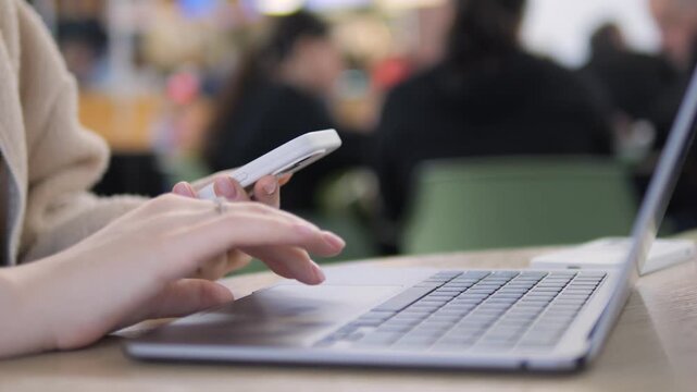 Close-up shows hands using smartphone and laptop. Person focused, multitasking in busy indoor setting. Ideal for tech, productivity, or communication themes