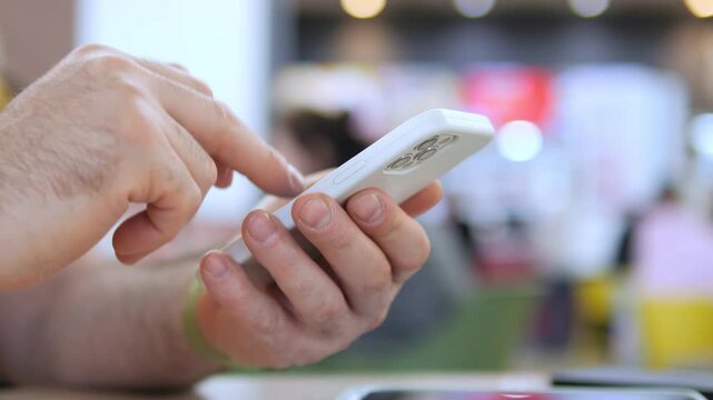Close-up of hands holding and tapping a white smartphone. Blurred background suggests public space. Person appears focused and engaged with device