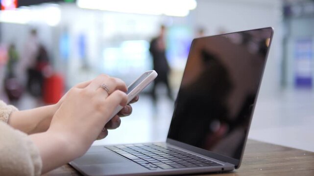 Person uses smartphone near open laptop. Hands hold phone, fingers touch screen. Background shows blurred indoor space with people moving. Laptop rests on wooden surface