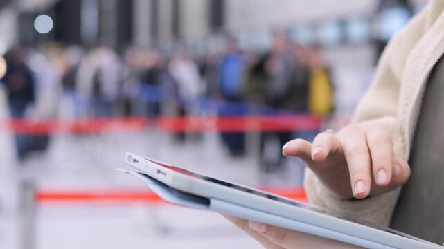 Hand holds tablet at airport terminal. Background shows blurred travelers and red stanchions. Focus on fingers tapping screen. No animals or plants visible
