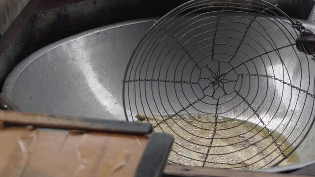Close up of deep frying traditional snacks or dough sticks in bubbling hot cooking oil inside a large metal wok with a wire mesh strainer skimmer spoon, authentic Asian street food cooking process. So