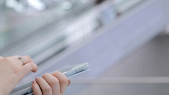 Person holds phone while riding escalator. Background shows blurred passengers moving upward. Modern indoor setting with reflective surfaces
