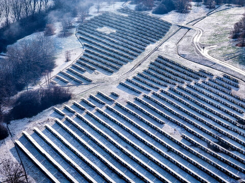 Aerial view of a solar park with rows of photovoltaic panels covered in light snow, surrounded by trees and rural landscape in Druento, Piedmont, Italy.