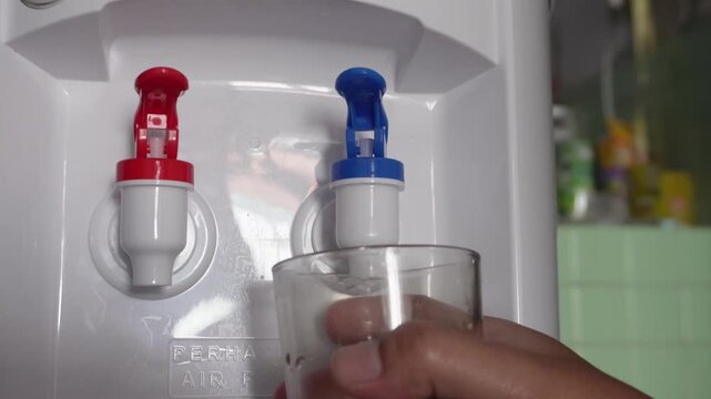 Close up of hand pressing a blue tap on a water dispenser machine to pour fresh cold water into a glass. Home or office appliance for domestic beverage refreshment