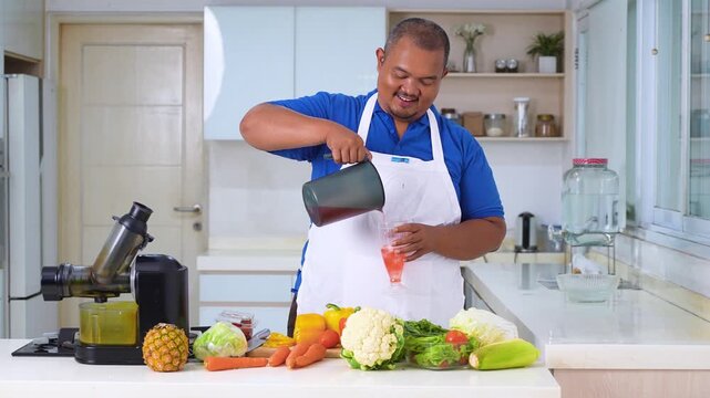 Smiling Asian man in a white apron pouring fresh vegetable juice into a glass in a bright kitchen.