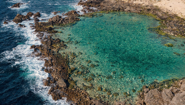 Aerial view of a natural turquoise rock pool on a rocky coastline with waves crashing against dark volcanic stones in the deep blue ocean water