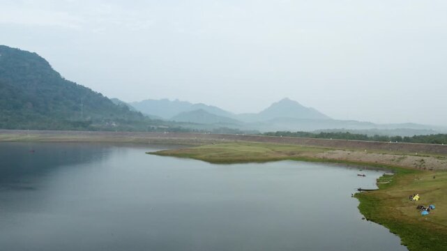 Scenic view of a wide lake reservoir or dam surrounded by mountains and hills under a hazy morning sky.