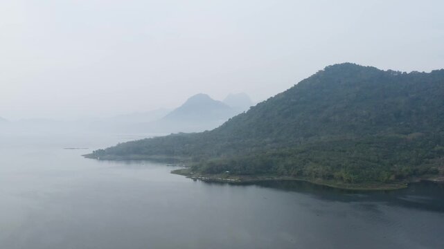 Beautiful aerial view of a large calm lake reservoir surrounded by lush green forested mountains under a misty sky.