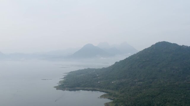 Stunning high-angle aerial view of a wide lake reservoir bordered by a lush green mountain ridge under a hazy sky.