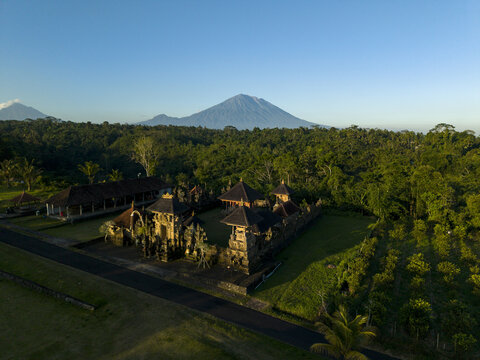 Aerial view of Pura Dalem Buungan temple complex surrounded by lush tropical forest and agricultural fields with Mount Agung in the background in Kecamatan Susut, Bali, Indonesia.