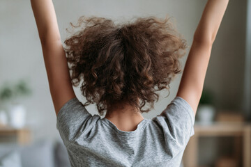 Fototapeta premium Curly-haired person stretches arms upward in quiet morning light. Back view shows relaxed posture, suggesting peaceful start to day. Soft gray shirt contrasts warm, natural tones of room