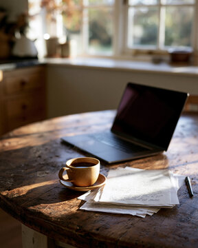 Warm sunlight glows on a rustic wooden table. Laptop rests beside a steaming coffee cup. Stacked papers and a pen suggest focused work or creative thought. The window frames a quiet