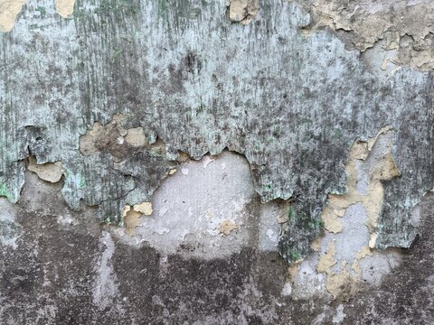 The rough, weathered texture of an old concrete wall. Its surface is uneven, featuring dark patches and mineral stains that reflect the natural aging process, along with peeling paint.