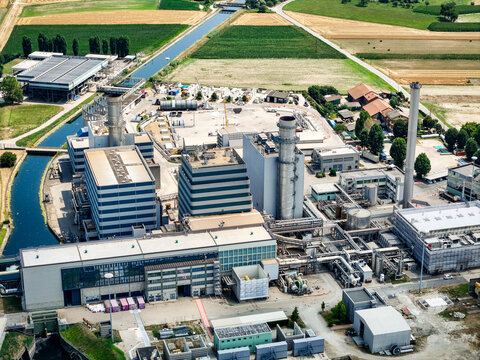 Aerial view of the Centrale Termoelettrica power plant with industrial buildings, tall chimneys, and piping systems near fields and a canal in Moncalieri, Piemonte, Italy.