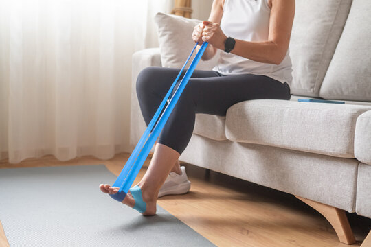 Woman sitting on a sofa performing foot rehabilitation exercises with a blue resistance band, focusing on recovery and strengthening muscles for physical therapy at home