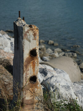 Rusted iron bars are attached to weathered concrete pillars along the seashore the exposed metal rebar is corroded by the salty air set against a backdrop of a green sea.