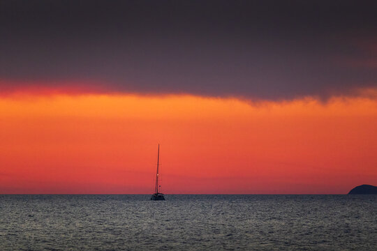 Solitary sailboat floating on the calm sea surface under a dramatic red and orange sunset sky with dark storm clouds, Crete
