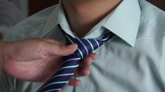 Close up of a woman's hand adjusting and fixing a blue striped necktie on a man's mint green shirt collar, preparing him for work, a wedding, or a formal event.
