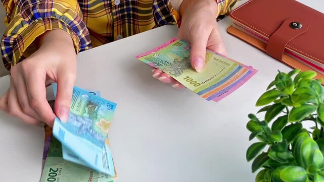 A close-up of a woman counting Indonesian rupiah banknotes at a table. Concepts for personal finance, budgeting, cash payments, travel, and money transactions.
