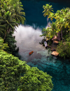 Aerial view of Danau Paisu Pok Luk Panenteng with a small boat on crystal clear blue water surrounded by lush palm trees and a wooden hut in Bulagi Utara, Central Sulawesi, Indonesia.