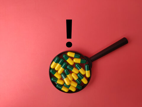 A black magnifying glass captures multiple green and yellow capsules beneath a black exclamation mark on red