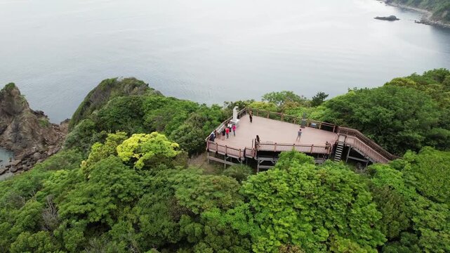 Aerial view of Lover's Cape coastal wooden deck surrounded by green trees, in Izu Peninsular Japan. People stand quietly, gazing at the sea from the viewpoint. Ocean waves crash against rocky shores b