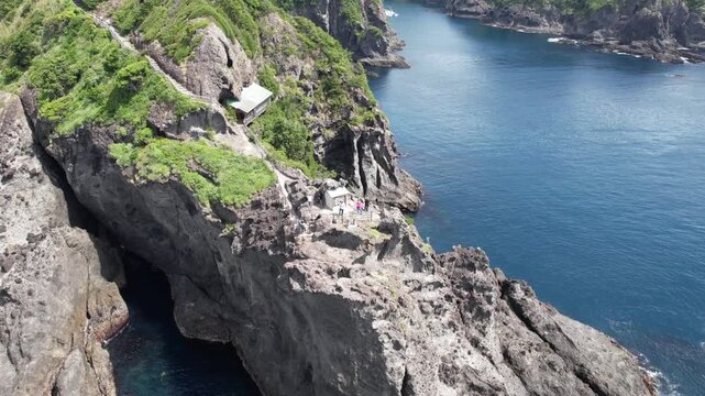 Japan's Izu Peninsular Cape Irozaki rugged cliffs overlook deep blue ocean waters. Greenery covers steep rocky terrain, adding vibrant contrast. Small structures dot the cliffside, offering shelter or