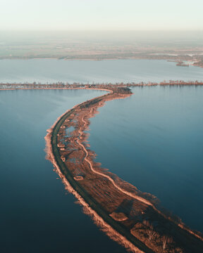 Aerial view of the narrow, curved Scheendijk land strip surrounded by calm waters, featuring grassy banks and a small road in Scheendijk, Breukelen, Utrecht, Netherlands.