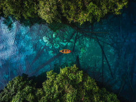 Aerial view of a wooden boat on the clear turquoise waters of Danau Paisu Pok Luk Panenteng, surrounded by lush forest and submerged logs in Bulagi Utara, Central Sulawesi, Indonesia.