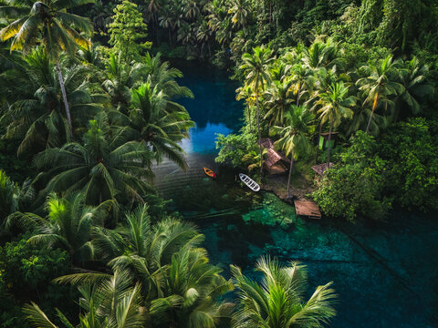 Aerial view of Danau Paisu Pok Luk Panenteng with crystal clear blue water surrounded by lush tropical palm trees and small wooden boats in Bulagi Utara, Central Sulawesi, Indonesia.