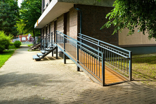 Metal wheelchair ramp with handrails leading to the entrance of a brick residential building on a sunny city street.
