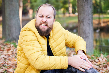 Fototapeta premium Calm explorer seated beside water body. Tranquil individual with beard waits amid colorful fallen leaves. Relaxed man sporting beard and yellow jacket sitting peacefully in autumn park setting