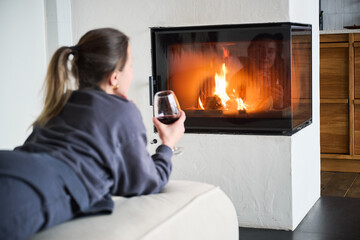Obraz premium Young woman resting on couch near burning fireplace, enjoying drink and calm atmosphere, cozy indoor leisure and relaxation scene